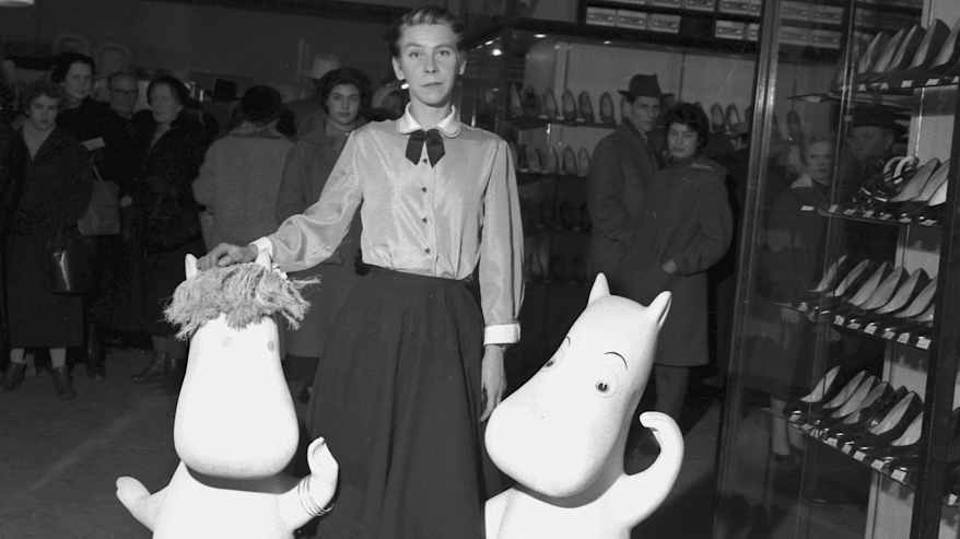 Mustavalkoisessa vanhassa kuvassa puseroon ja hameeseen pukeutunut henkilö poseeraa kahden muumihahmon kanssa tavaratalossa, taustalla ihmisiä.