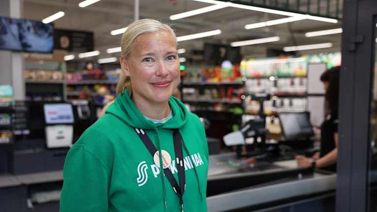 Nainen S-kaupan vihreässä hupparissa katsoo kohti ja hymyilee.