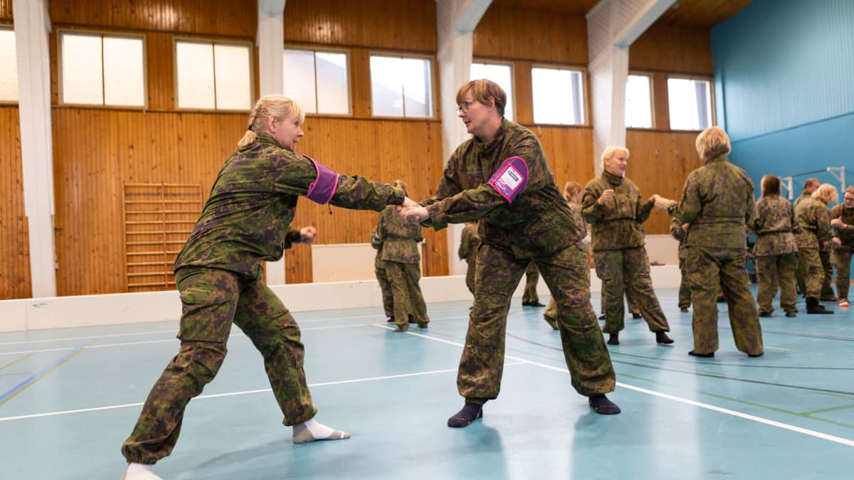 Maastopukuiset naiset harjoittelevat itsepuolustusotteita liikuntasalissa.