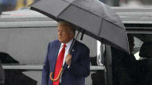 Ulkona sataa. Trump seisoo auton vieressä ja pitelee sateenvarjoa yllään.