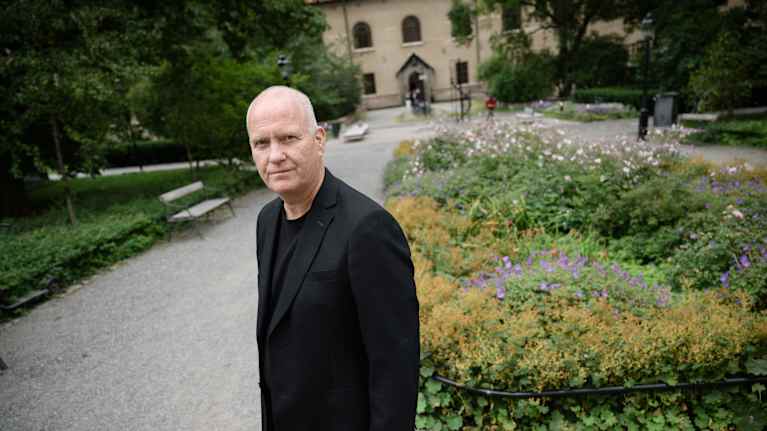Henkilö seisoo puistossa, joka on täynnä vehreää kasvillisuutta ja kukkia. Taustalla näkyy vanha rakennus.