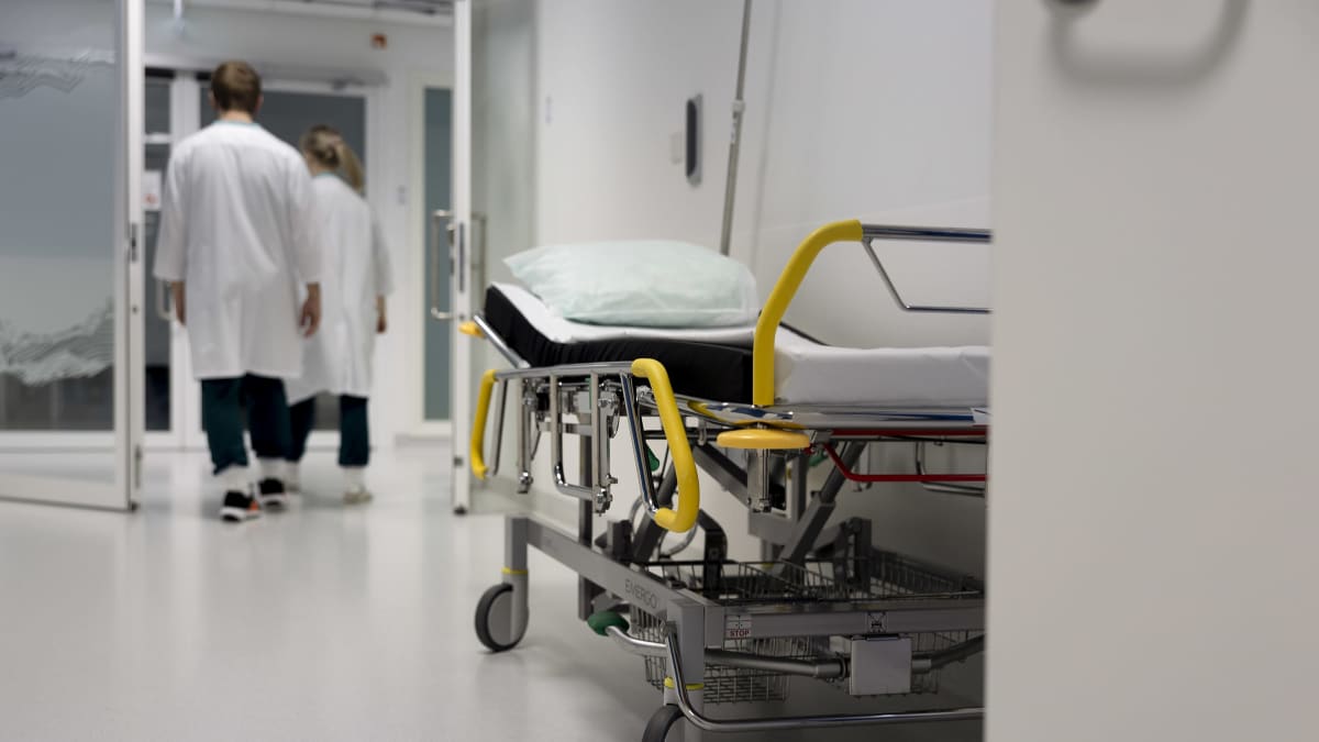 Lääkärit kävelevät Keski-Suomen keskussairaala Novan päivystyksen käytävällä. Käytävän seinustalla seisoo tyhjä potilassänky. 