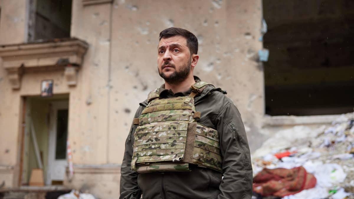 Volodymyr Zelenskyi seisoo vakavailmeisenä sodan tuhoaman rakennuksen edessä.