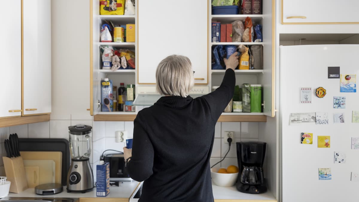 Anne Matilainen pakkaa keittiön kaappeihin ruoka-ainepakkauksia. Hänen selkänsä on kohti kameraa. 