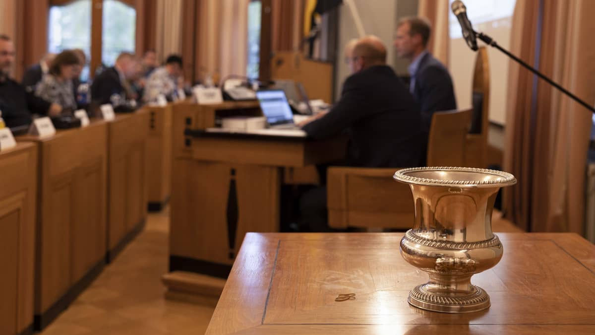 Mikkelin kaupunginjohtajan valinnassa käytetty kiiltävä metalliastia seisoo puupöydällä Mikkelin kaupunginvaltuuston salissa. Taustalla siintää valtuutettuja.