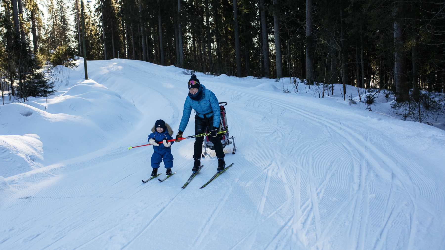 Susanna Ylinen laskee pienen lapsensa kanssa hiihtosuksilla mäkeä alas. Lapsi pitää kiinni Susannan sauvoista.