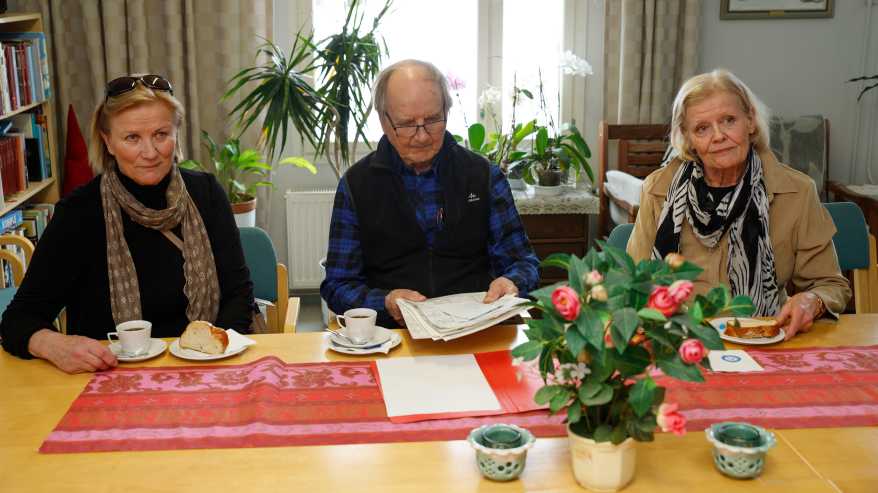 Marja Puhakka, Ismo Mikkonen ja Paula Lempiäinen istuvat pöydän ääressä. Edessään heillä on kahvikupit ja pullaa.