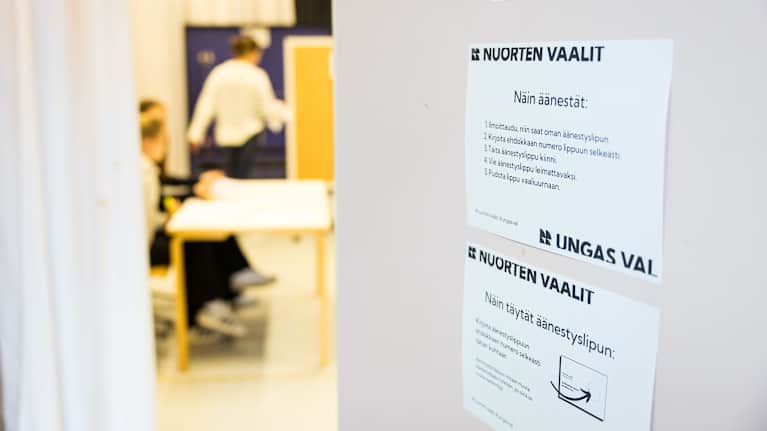 Nuorten vaalit äänestysohjeet monisteissa oveen kiinnitettynä. Taustalla äänestyspiste, missä vaalivalvojat istuvat pöydän takana.