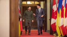 Zelenskyi ja Trudeau kävelevät vierekkäin käytävällä, jota reunustavat lippurivit.