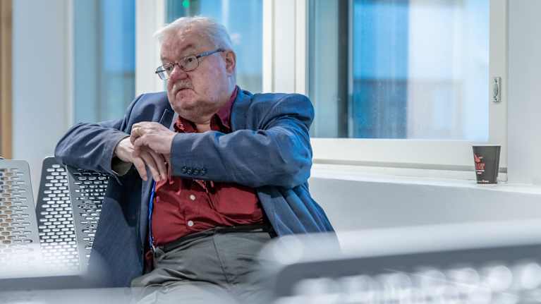 Elderly man in blue blazer and red shirt sitting in a chair, glancing to his right.