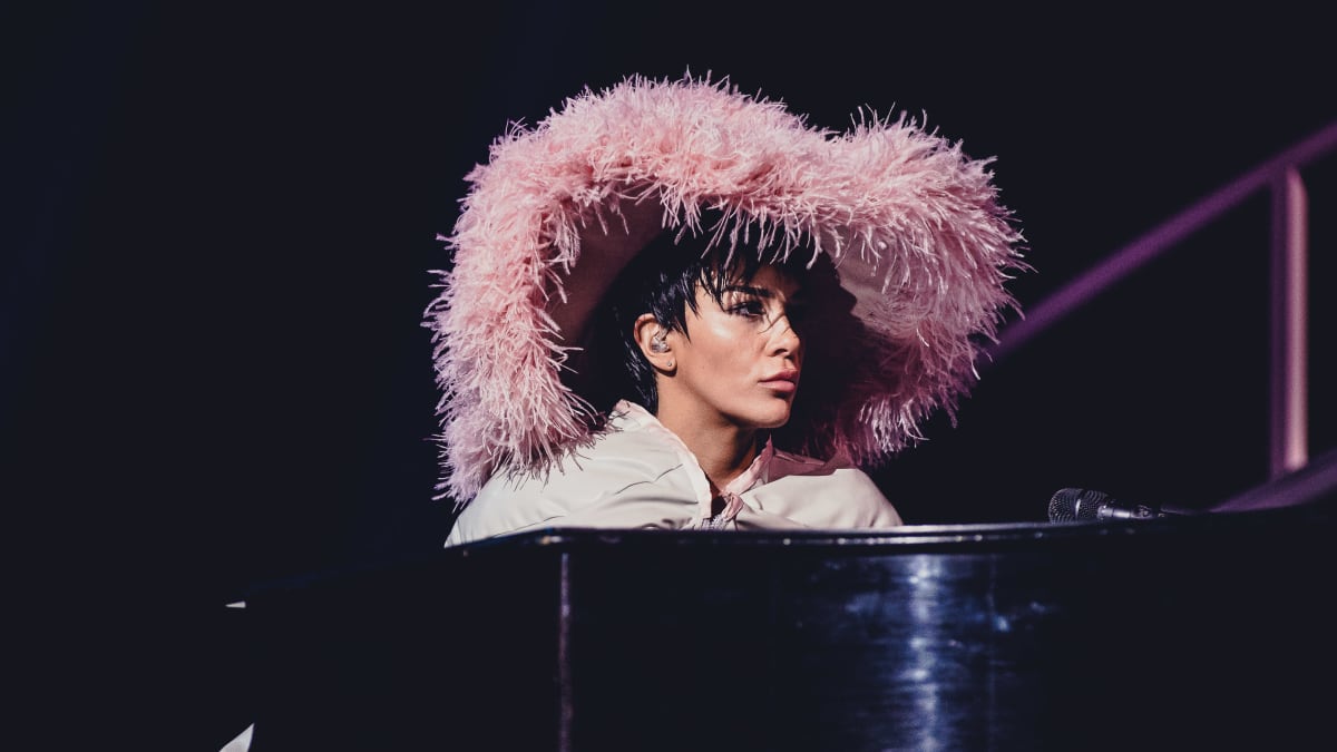Massiiviseen vaaleanpunaiseen höyhenpäähineeseen pukeutunut Sini Sabotage istumassa mustan pianon äärellä.