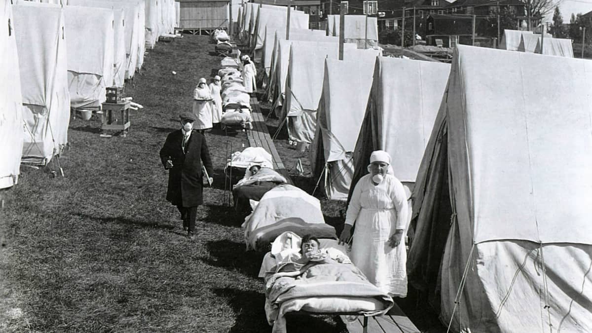 Kentällä on rivi telttoja ja telttojen edessä kenttävuoteilla makaa sairaita. Heidän vieressään seisoo sairaanhoitajia. Tummapukuinen mies kulkee telttojen välissä kulkevalla polulla.