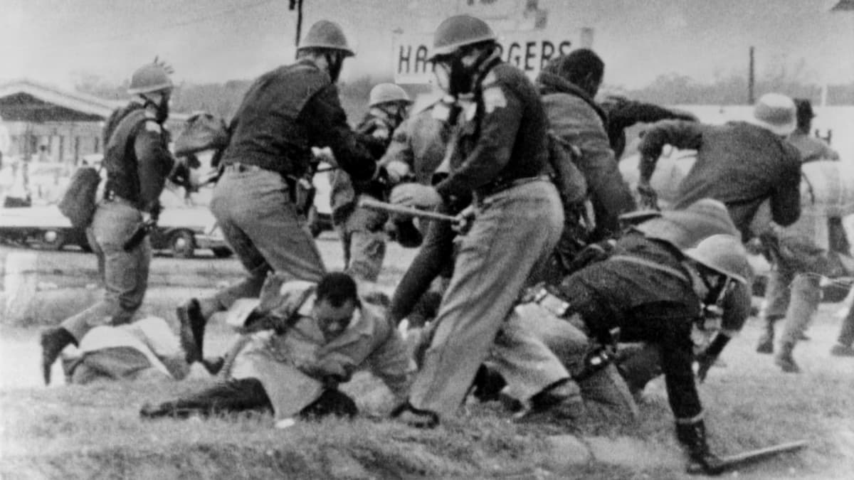Mustavalkoisessa kuvassa valkoiset kypäräpäiset poliisit hakkaavat pampuilla maassa makaavia mustia. 