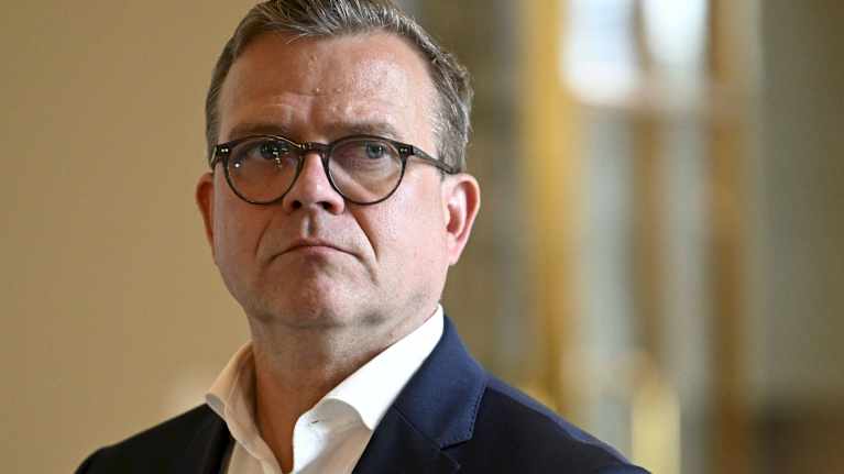 Close-up of a man wearing round horn-rimmed glasses, a serious expression, an open-necked shirt and a dark blue suit jacket.  