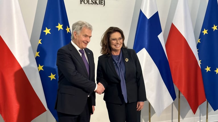 Harmaantunut pukumies ja tumma pukunainen hymyilevät levästi oikealle kameroille. Taustalla Puolan valkopunaisia, Suomen sinivalkoisia ja EU:n keltasinisiä lippuja.