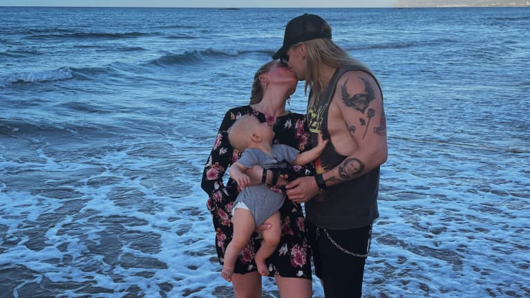 Olli Herman suutelee etelänlomalla rantavedessä puolisoaan Tyttiä, jolla on sylissä heidän lapsensa.