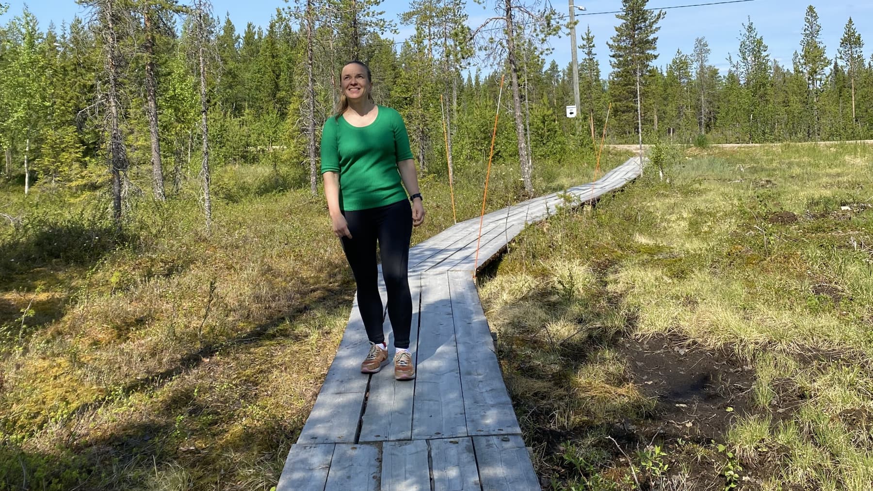 Kirjailija Satu Rämö kävelee pitkospuilla Äkäslompolossa, katse on suunnattuna aurinkoiselle taivaalle. Maisema on kesäinen.