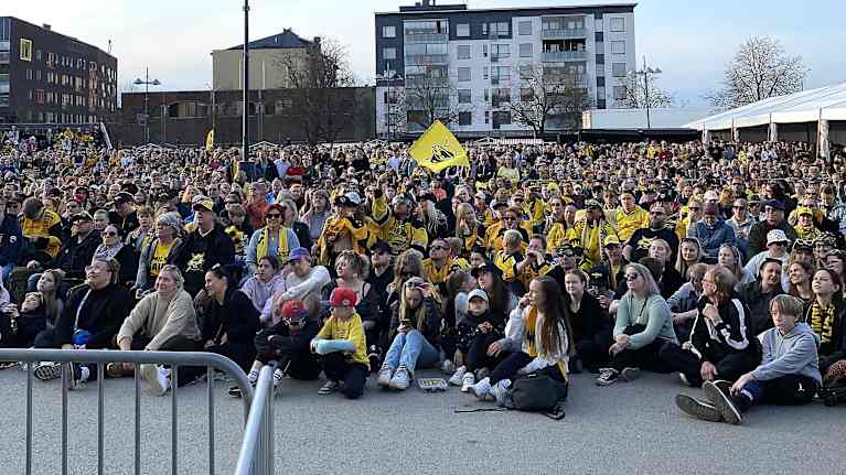 Tuhansia ihmisiä istuu asvaltilla, osa seisoo. Osalla Saipa-asu päällä. Taustalla kauppatori. 