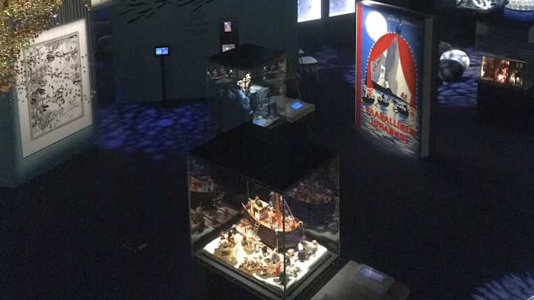 Näkymä Muumimuseon ylätasanteelta alas. Etualalla on pyrstötähteä esittävä​ helmiteos ja alhaalla näkyy pienoismaailmoja‍ lasikuvuissaan.