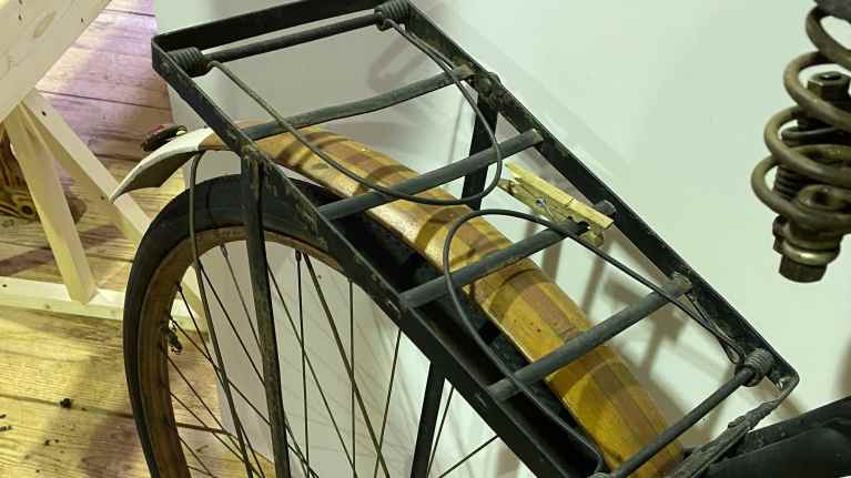 Polkupyörän takaosa: metallinen tarakka, johon on kiinnitetty pyykkipoika, vaaleankeltainen, puusta valmistettu takavanne, jossa keskellä ruskea raita. Ruskea, puusta valmistettu vanne kiertää takapyörää.