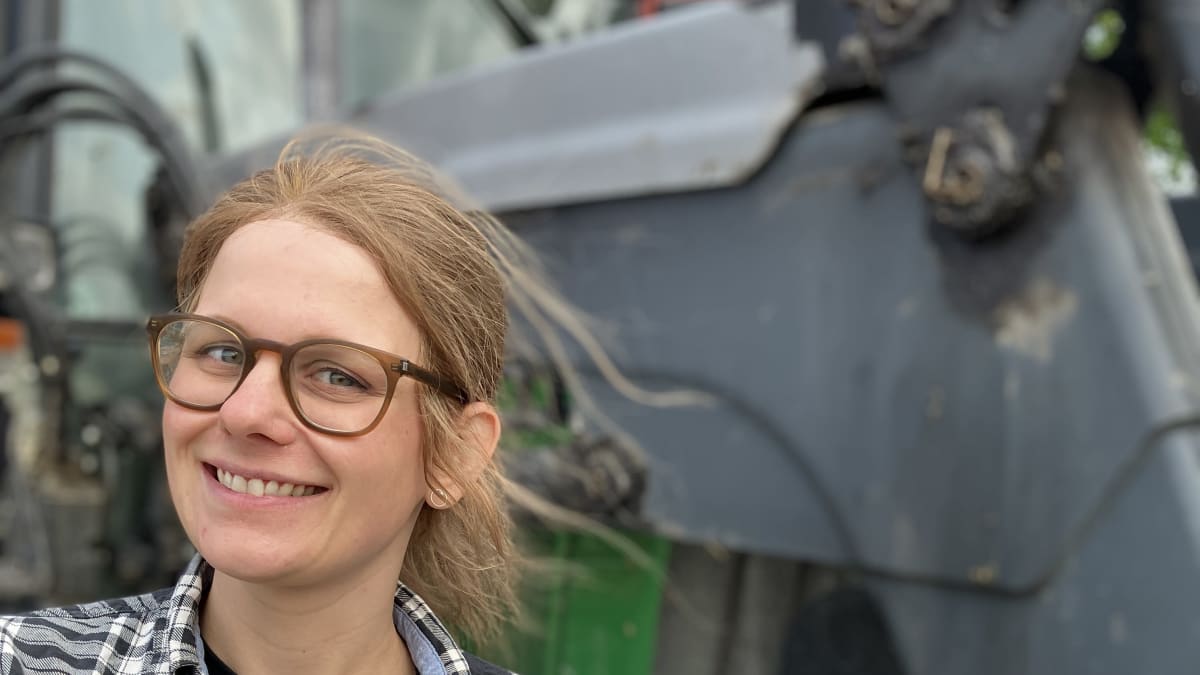 Silmälasipäinen nainen poseeraa traktorin edessä ja hymyilee kameralle.