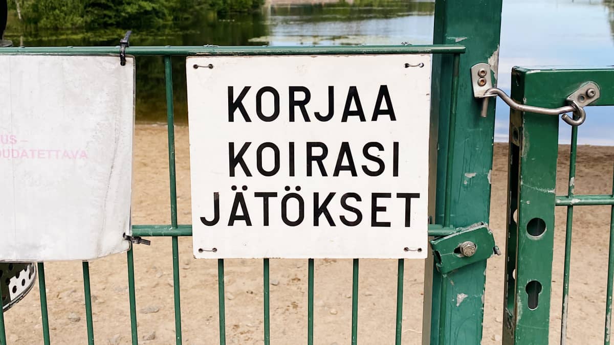 Korjaa koirasi jätökset -kyltti koirauimarannan aidassa. Taustalla Saimaa.