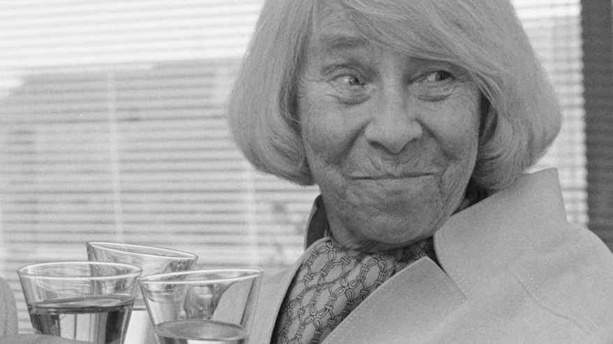 Mustavalkoisessa valokuvassa valkohiuksinen henkilö hymyilee kuohuviinilasi kädessä, kuvassa näkyy laseja myös muiden henkilöiden käsissä.