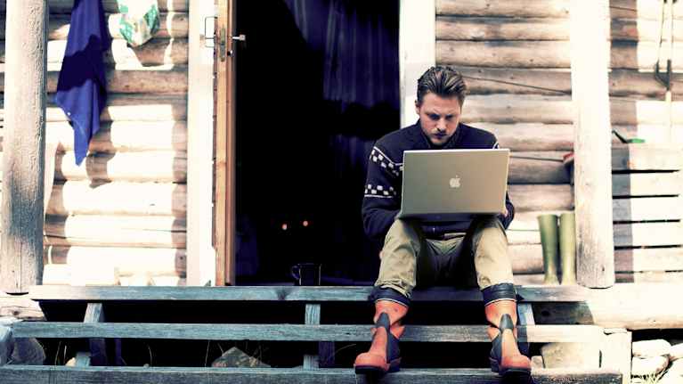 A man wearing rubber boots and a wool jumper sits working at a laptop on the steps of a rustic cabin.