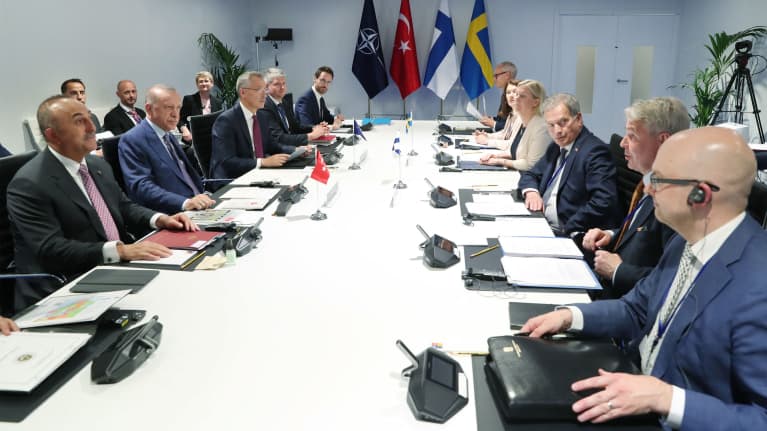 Pitkän pöydän ääressä ihmisiä. Taustalla Naton, Turkin, Suomen ja Ruotsin liput