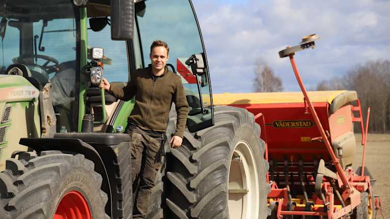 En person står bredvid en traktor, med en såmaskin kopplad bakom. Soligt väder med en molnig himmel.