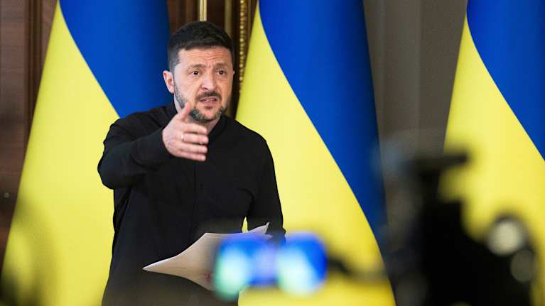 Volodymyr Zelenskyi speaks in front of Ukrainian flags.