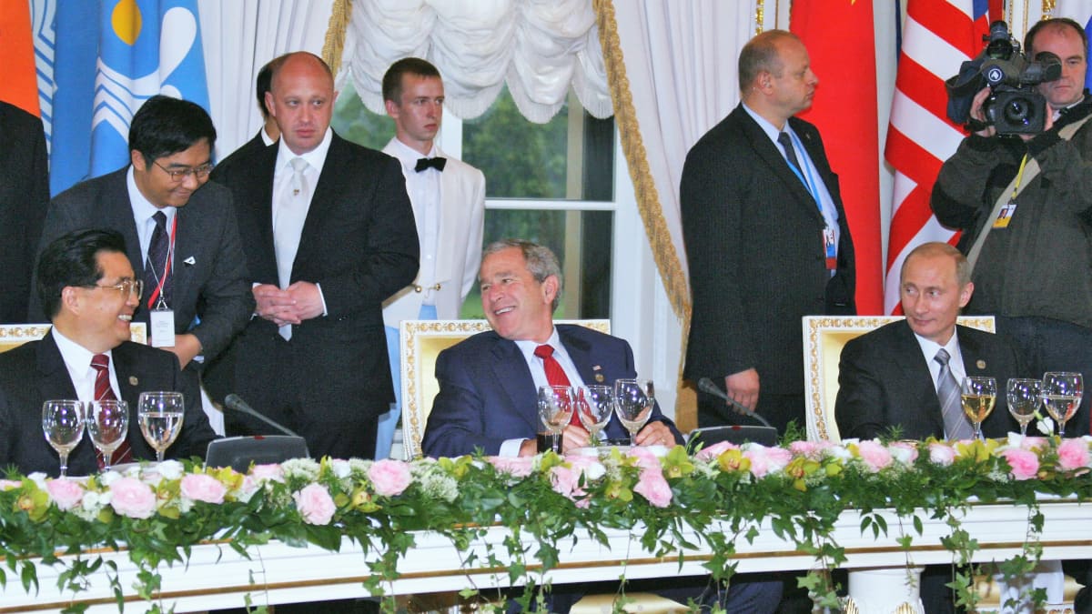 Hu Jintao, George Bush nuorempi ja Vladimir Putin istuvat tummissa puvuissa pöydän takana. Hu ja Bush nauravat, Putin hymyilee. Taustalla Hun ja Bushin välissä näkyy Prigožin mustassa puvussa ja vaaleassa kravatissa.