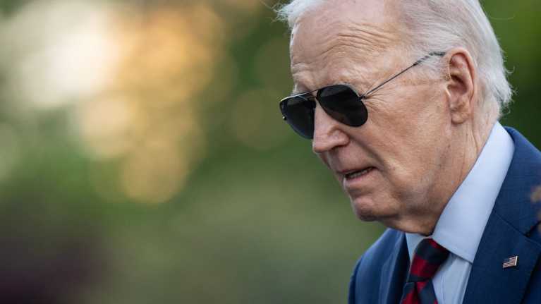 Joe Biden i en mörkblå kostym med slips tittar åt sidan och har på sig solglasögon.