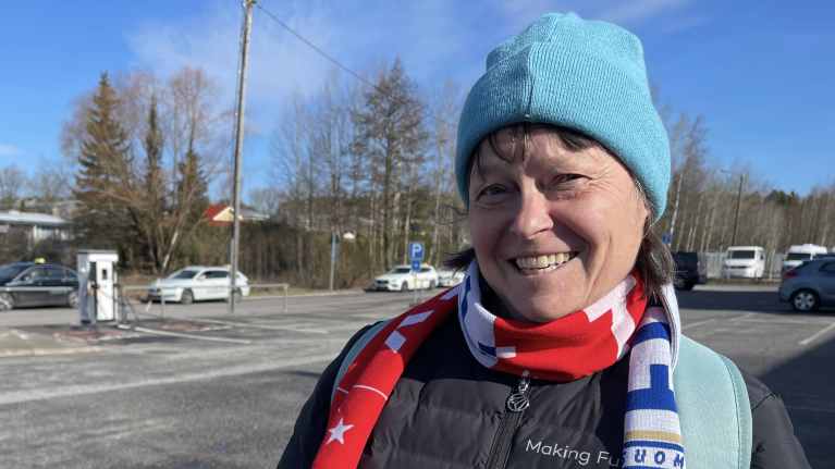 Nainen hymyilee Suomi-kaulahuivi yllään, taustalla autoja ja parkkipaikka.