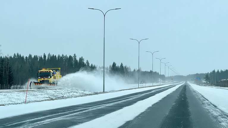 Keltainen aura-auto ajaa moottoritiellä ja lumi pöllyää.