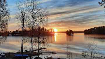 Kaunis auringonlasku järvimaisemassa, jossa taivaanranta hehkuu oranssin ja sinisen sävyissä, heijastuen tyynestä vedestä.