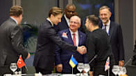 Six men standing in a conference hall, all but one wearing dark suits and two of them shaking hands. 