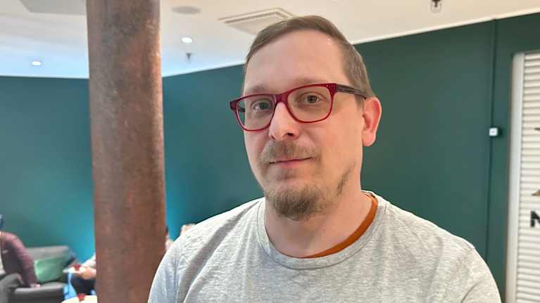 En person med grå tröja står i ett rum med gröna väggar, där flera andra människor också syns i bakgrunden.
