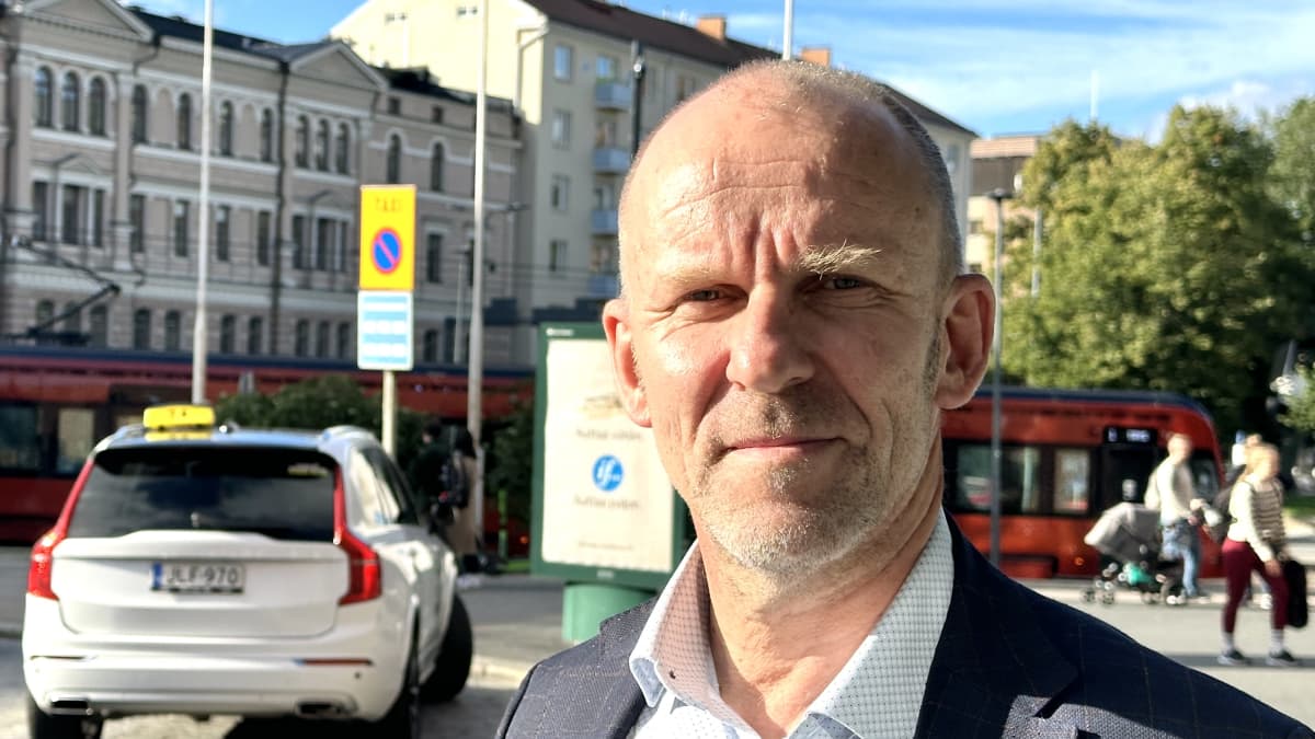Petri Räsänen seisoo Tampereen keskustassa rautatieaseman edessä. Kuvassa näkyy taustalla taksi, ratikka ja ihmisiä.