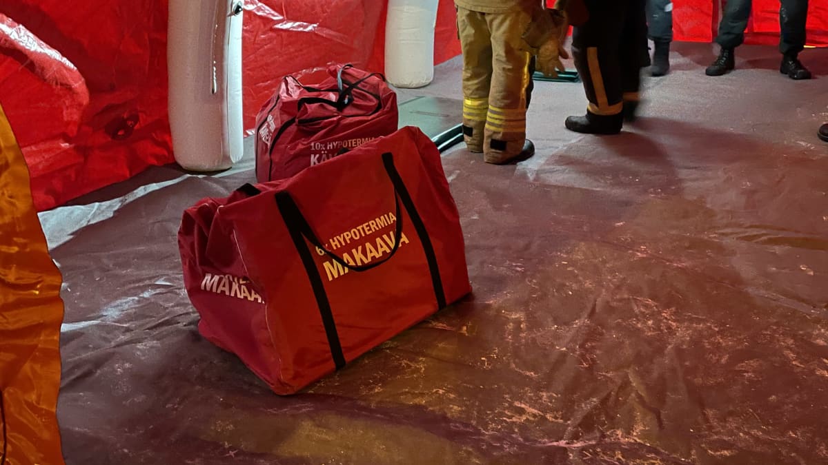 Punaisen ilmatäytteisen teltan sisällä on punainen iso kassi, jossa teksti hypotermia makaava. Taustalla virakapukuisia ihmisiä.