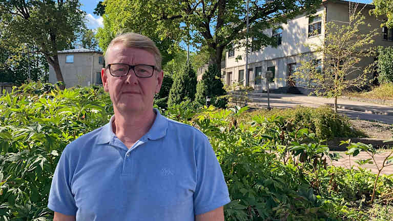 En man med svarta glasögon, blont hår och ljusblå pikétröja står framför ett buskage.