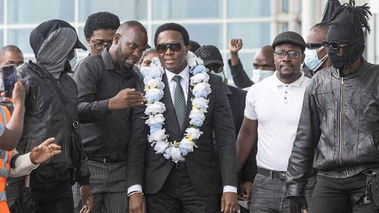 Venancio Mondlane (keskellä) mustassa puvussa, sinivalkoinen kukka seppele kaulalla. Ympärillä tummaan pukeutuneita miehiä. Osalla kasvomaskit.