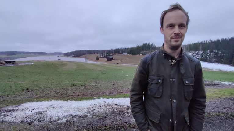 En man står framför en åker med höstvete och Raseborgs å som svämmar över. Han heter Thomas Björklöf.