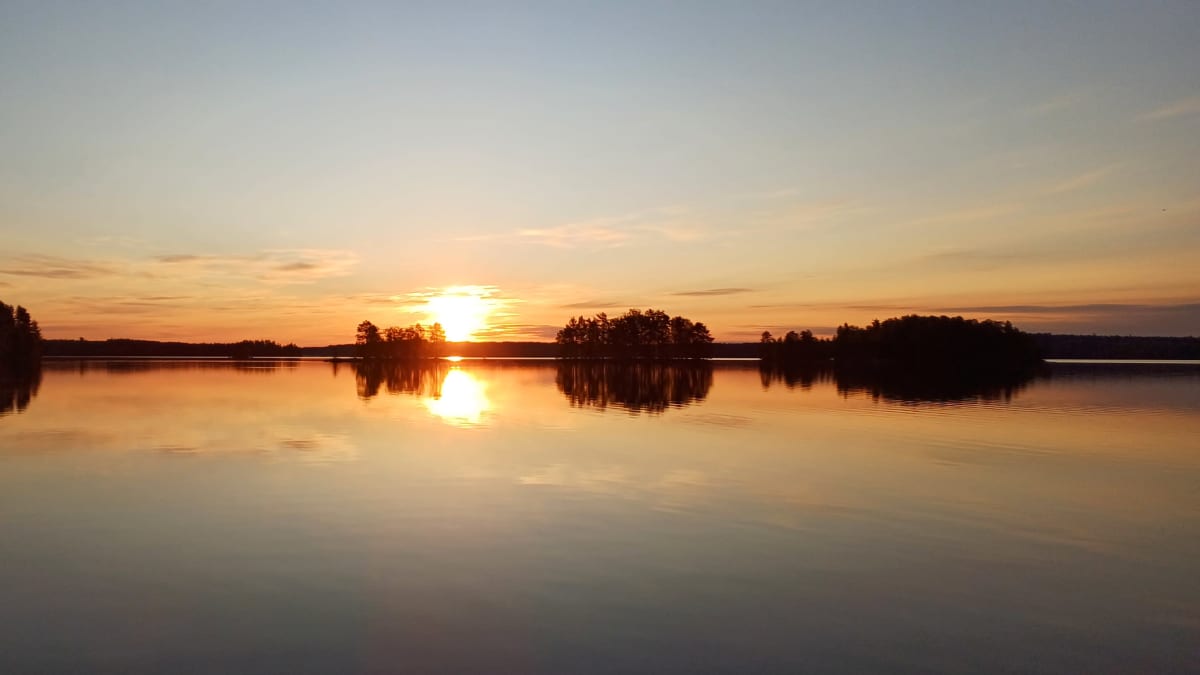 Auringonnousu Kiteellä. Aurinko nousee järven takaa, tyyni sää ja saaria. 