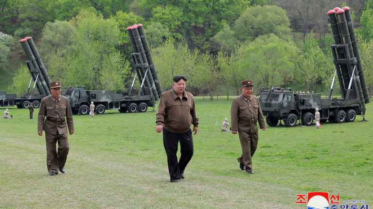 Kim Jong-un ja kaksi sotilashenkilöä kävelee nurmikentällä. Taustalla ohjusjärjestelmiä.