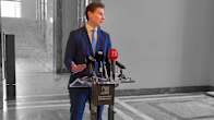 A man in a blue suit stands speaking in a corridor at a small podium with microphones and the Parliament logo and name in Finnish and Swedish.
