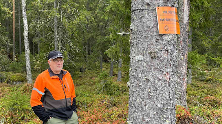 En man i orange och svart jacka står i skog bredvid ett träd med en skylt där det står "MILJÖSKYDD"