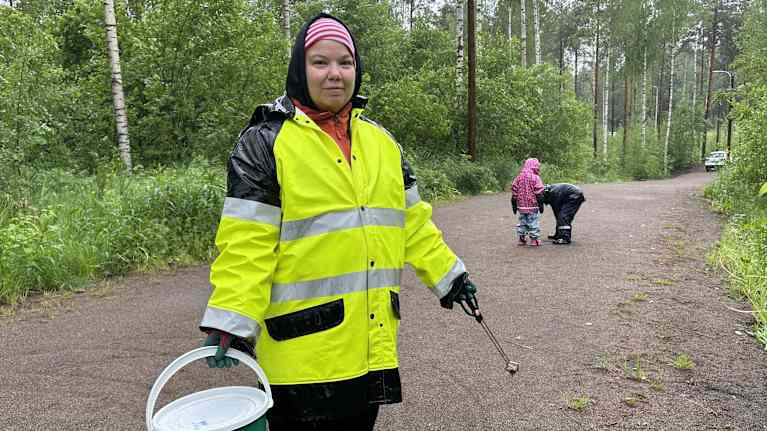 Henkilö kerää etanoita metsäisessä ympäristössä, pukeutuneena kirkkaan keltaiseen sadeasuun. Taustalla lapset tutkivat tieltä löytynyttä kastematoa.