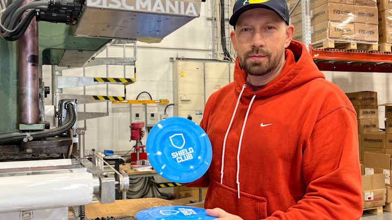 Jussi Meresmaa katsoo kohti kameraa. Hän pitää sinisiä frisbeegolf-kiekkoja käsillään, taustalla näkyy varastotiloja.