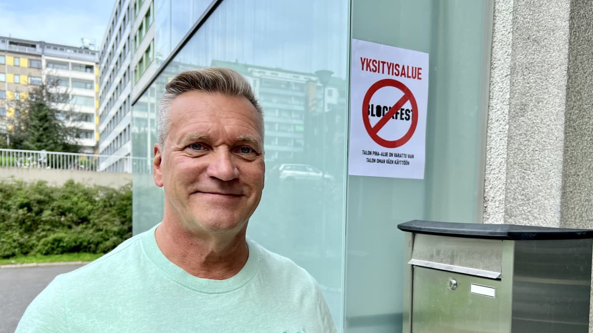 Mies seisoo kerrostalon edessä ja hymyilee kameralle. Talon seinässä on kyltti, jonka teksti kertoo talon piha-alueen olevan varattu vain talon oman väen käyttöön.